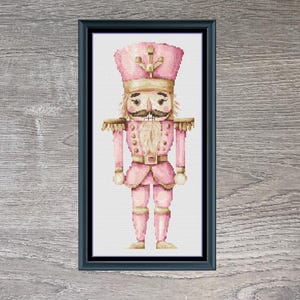 May include: A framed cross-stitch of a pink Christmas soldier nutcracker. The artwork is in a black frame and is set against a wood-grain background. The text "Little Christmas Soldier" is below the framed art. The dimensions are 90 x 200.