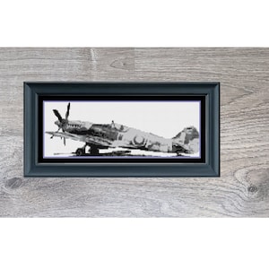 May include: A framed black and white cross-stitch of a mini fighter plane. The artwork is in a black frame, set against a gray wooden background. The text "Mini Fighter Plane" is below the frame.
