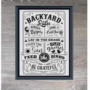 May include: Framed black and white cross-stitch art with the text "Backyard Rules". The design includes phrases like "Grill Burgers", "Drink a Cold One", and "Feed the Birds". The frame is dark gray and the dimensions are 254 x 349.