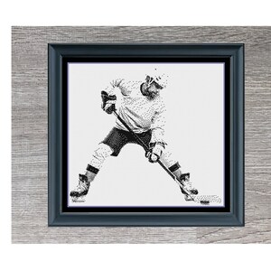 Könnte beinhalten: Schwarzweißes Kreuzstichmuster eines Hockeyspielers in Aktion, gerahmt in einem grauen Holzrahmen mit schwarzem Passepartout. Der Titel "Hockey Player" ist unter dem Rahmen gedruckt.