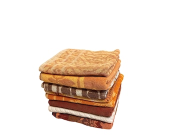 May include: A stack of seven folded terry cloth towels in various shades of orange, brown, and cream. Each towel features a distinct woven texture or pattern, including geometric shapes, stripes, and floral motifs, creating a layered appearance.