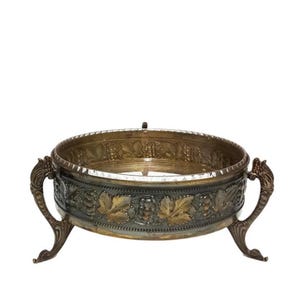 May include: A round, silver-toned serving dish with three legs and ornate, gold-toned grapevine designs around the rim.