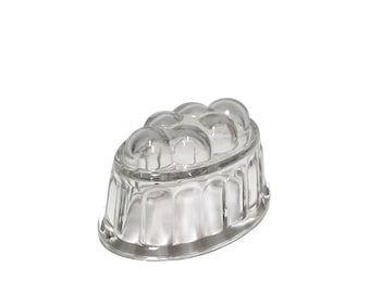 May include: A clear glass jelly mold with a rounded top and a scalloped base. The mold has a series of rounded indentations on the top surface. The glass is transparent, allowing light to pass through. The mold is designed for making molded desserts.