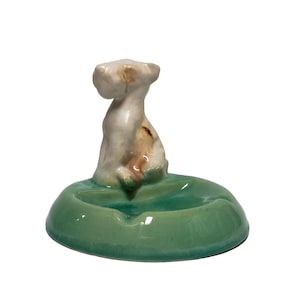 May include: A ceramic ashtray with a white dog figurine on top. The ashtray is green and has a round shape.