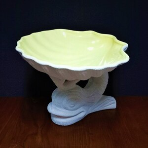 May include: A white ceramic bowl shaped like a seashell sits on a white ceramic dolphin figurine. The bowl is yellow on the inside.