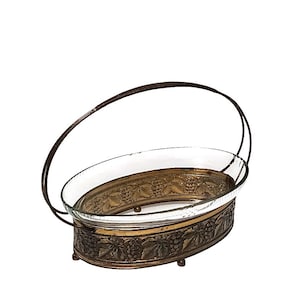 May include: An oval serving dish with a clear glass insert and a decorative metal frame. The frame features a grape vine design and a handle. The dish is supported by small feet and is suitable for serving food.