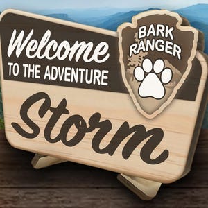 May include: Wooden sign with the text "Welcome to the Adventure" and the name "Storm". A "Bark Ranger" badge with a paw print is attached. The background shows a mountain landscape.