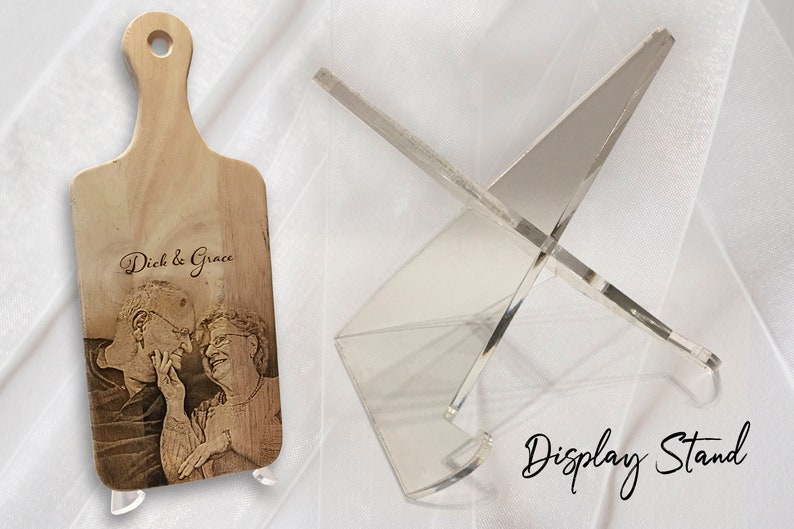 May include: A personalised wooden chopping board with a laser engraved photo of a couple and the text "Dick & Grace". The board is displayed on a clear acrylic stand.