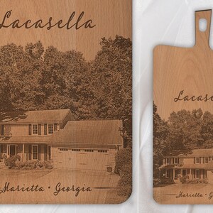 May include: A personalised wooden chopping board with a laser engraved image of a house and trees. The text "Lacasella" and "Marietta, Georgia" is engraved on the board. The board is made of oblique beech wood.