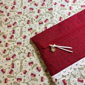 Puede incluir: Una bolsa roja con cremallera, estampado floral y ribete de encaje blanco. El tirador de la cremallera tiene un dije de margarita. El fondo es una tela con estampado floral.