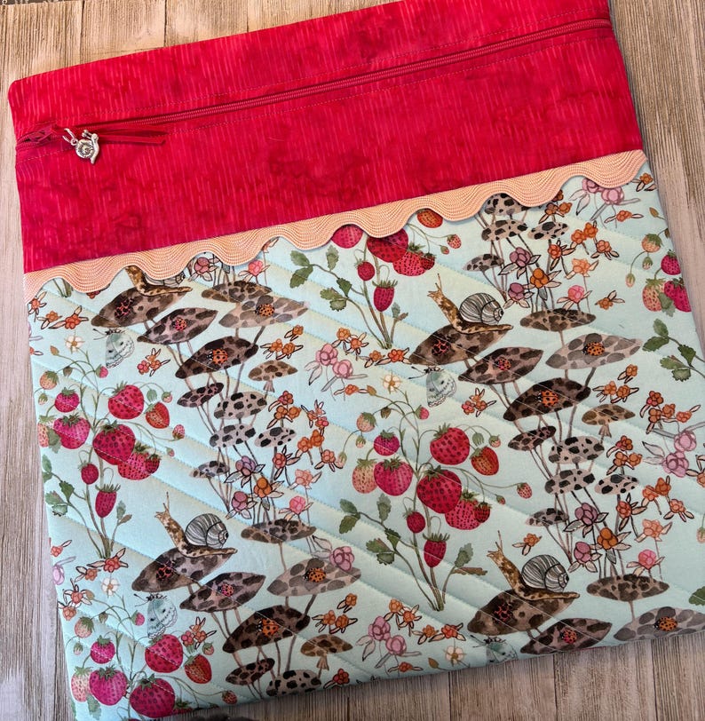 Pu&ograve; includere: Una borsa in tessuto trapuntato con parte superiore rossa e corpo azzurro. Il corpo presenta una stampa stravagante di fragole, lumache e funghi. Un bordo smerlato separa la parte superiore e il corpo. La borsa ha una chiusura a zip e un ciondolo decorativo.