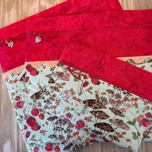 Pu&ograve; includere: Tre pochette con cerniera trapuntate con sfondo azzurro chiaro con un motivo a fragole e lumache. La parte superiore di ogni pochette &egrave; di un rosso vibrante con una cerniera abbinata. Ogni tiretto della cerniera ha un ciondolo decorativo.