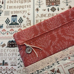 May include: A rectangular zippered pouch featuring a coral-colored fabric with a white botanical print, accented by a cream-colored lace trim and a small circular charm on the zipper pull, resting on a white fabric with cross-stitch patterns.