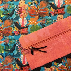 May include: A rectangular coral-colored zippered pouch featuring a black bird charm on the zipper pull and a black scalloped trim along the bottom edge. The pouch is placed against a fabric background with a teal pattern of black birds and foliage.