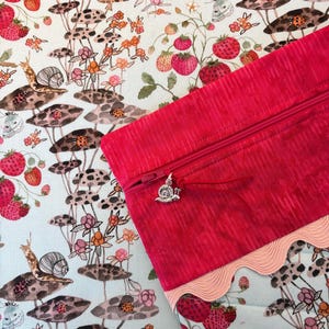 Pu&ograve; includere: Una pochette con cerniera lampo rosa acceso con un ciondolo a forma di lumaca, ambientata su un tessuto stravagante con fragole, funghi, lumache e coccinelle. Il tessuto ha uno sfondo azzurro con un bordo smerlato.