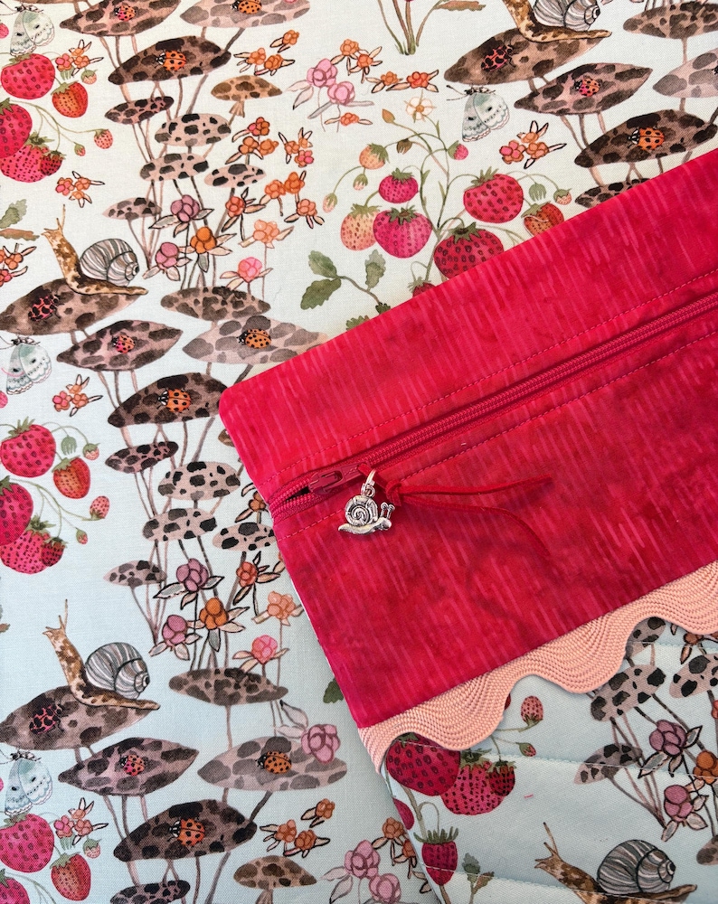 Pu&ograve; includere: Una pochette con cerniera lampo rosa acceso con un ciondolo a forma di lumaca, ambientata su un tessuto stravagante con fragole, funghi, lumache e coccinelle. Il tessuto ha uno sfondo azzurro con un bordo smerlato.