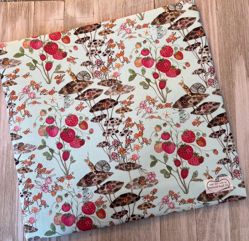 Pu&ograve; includere: Un tessuto azzurro con un motivo stravagante di fragole e funghi. Il design presenta fragole rosse, funghi marroni, lumache e piccoli fiori. Il tessuto ha un'etichetta che dice "made with love by Painted Leaf Co."