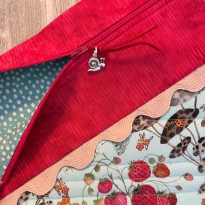 Pu&ograve; includere: Primo piano di una pochette rossa con cerniera e un ciondolo a forma di lumaca argentata. La pochette ha una fodera a pois azzurri e un bordo decorativo. Il tessuto inferiore presenta un motivo a fragole e funghi.