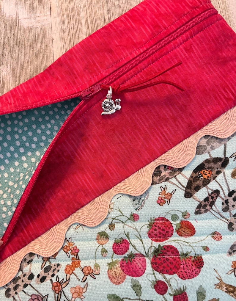 Pu&ograve; includere: Primo piano di una pochette rossa con cerniera e un ciondolo a forma di lumaca argentata. La pochette ha una fodera a pois azzurri e un bordo decorativo. Il tessuto inferiore presenta un motivo a fragole e funghi.
