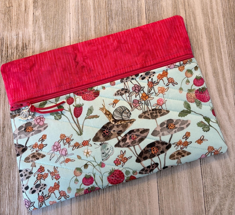 Pu&ograve; includere: Pochette in tessuto trapuntato con sfondo azzurro chiaro e fantasia di fragole e funghi. La parte superiore &egrave; rossa a tinta unita con chiusura a zip e un ciondolo decorativo. Ideale per trasportare piccoli oggetti.