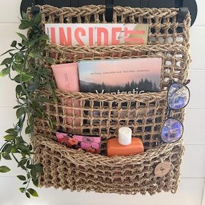 May include: A woven jute wall organiser with three pockets, holding magazines, a pink tube, a small cosmetic bag, and an orange bottle. The organiser has a black top mount and is decorated with a green vine and a wooden tag.