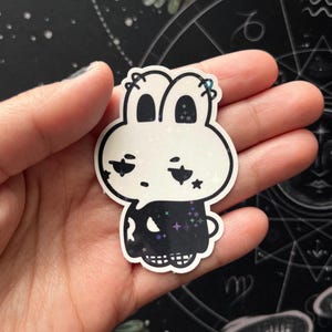 May include: A white and black sticker featuring a cartoon bunny with a skull-like design on its lower half. The bunny has star accents and a glittery finish. The sticker is held in a hand against a dark background with astrological symbols.