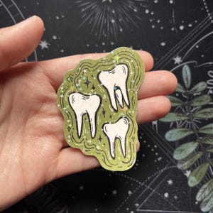 May include: A sticker featuring three white teeth with black outlines on a green background. The teeth are surrounded by a sparkly design with stars. The sticker is held in a hand.