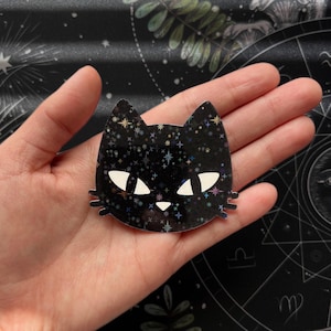 May include: A black cat head sticker with a starry holographic design. The cat's face has white eyes and a white nose. The sticker is held in a hand against a dark background.