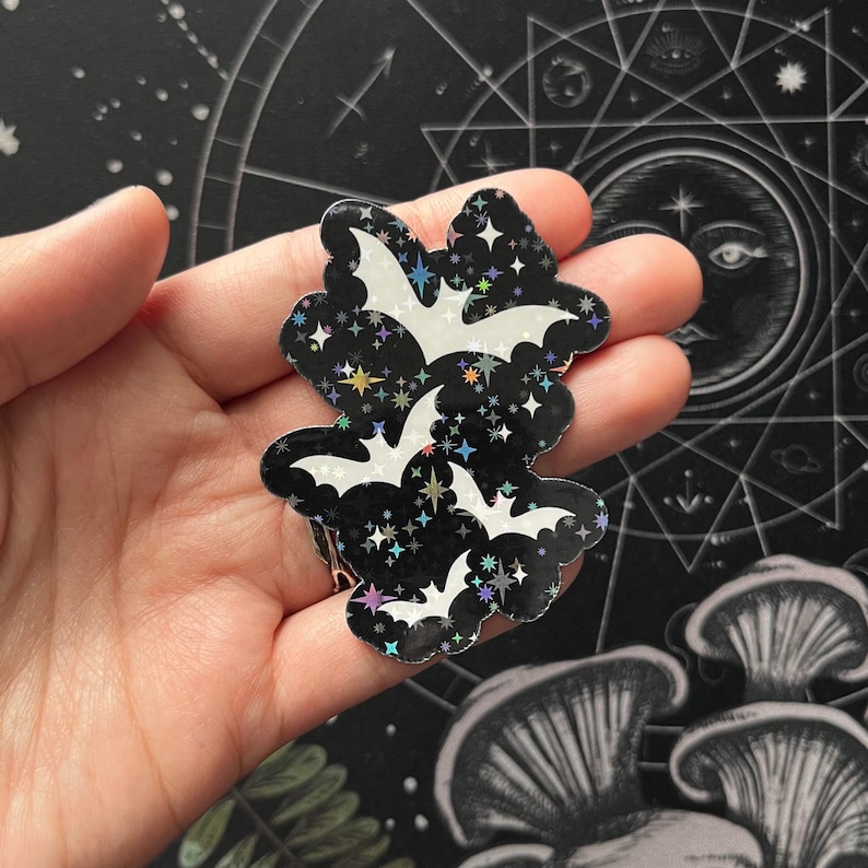 May include: A black and iridescent bat-shaped sticker with three white bat silhouettes and star details. The sticker is held in a hand, with a dark background featuring celestial and mushroom illustrations.