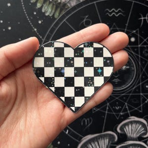 May include: A heart-shaped sticker with a black and white checkered pattern. The sticker has a holographic finish with small, colorful star accents. The sticker is held in a hand against a dark background with astrological symbols.
