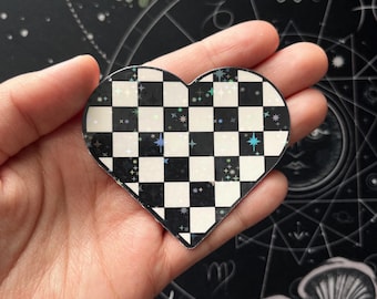 May include: A heart-shaped sticker with a black and white checkered pattern. The sticker has a holographic finish with small, colorful star accents. The sticker is held in a hand against a dark background with astrological symbols.