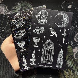 May include: Two black sticker sheets with white illustrations of gothic and medieval elements. One sheet has a holographic, starry background. The stickers include a helmet, books, chalice, candle, raven, dagger, and a moon.