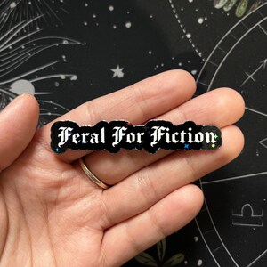 May include: A black rectangular pin with the text "Feral For Fiction" in white gothic lettering. The pin has a glittery, iridescent border with small blue and yellow star accents. The pin is held in a hand.