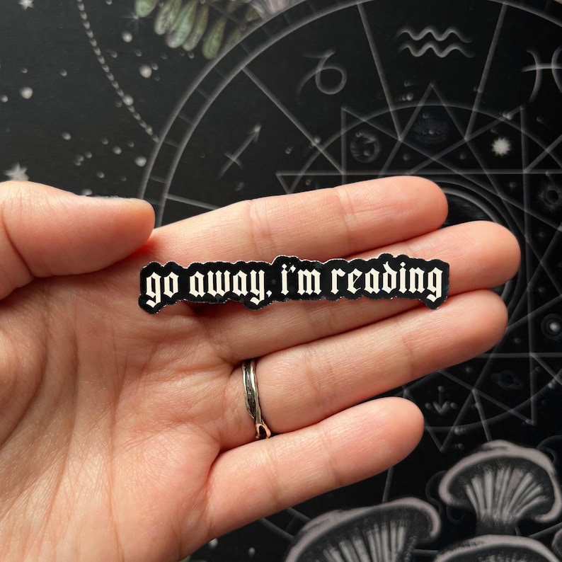 May include: A black and white rectangular pin with the text "go away, I'm reading" in a gothic font. The pin is held in a hand, with a silver ring on the finger. The background is a dark surface with astrological symbols.