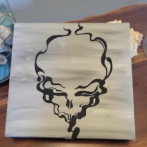 Puede incluir: Arte en lienzo cuadrado con un diseño de calavera abstracta negra sobre un fondo gris y blanco. La calavera está representada con líneas fluidas y estilizadas, creando una representación artística única. La obra de arte está montada en un marco de madera.