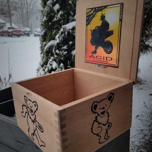 Puede incluir: Una caja de madera con una tapa con bisagras. La tapa presenta un gráfico de una persona conduciendo una motocicleta con el texto "ACID CIGARS BY DRAW ESTATE" y "MANTRA". La caja tiene dos gráficos de osos negros y blancos en los lados.