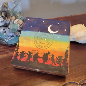 Puede incluir: Caja de madera pintada a mano con un diseño de cielo nocturno. La caja presenta una luna creciente, estrellas y una silueta de osos bailando. La caja está pintada con un degradado de colores, incluyendo azul, amarillo y naranja.
