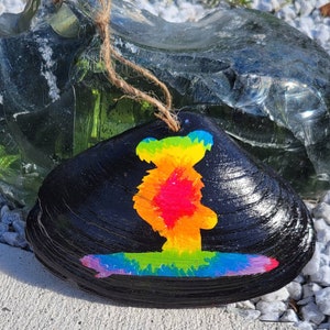 May include: A black seashell painted with a tie-dye design of a bear surfing on a rainbow wave.