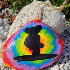 May include: A colorful tie-dye painted seashell ornament with a black silhouette of a bear surfing on a wave.