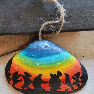 May include: Hand-painted seashell ornament featuring a colorful rainbow sky with a crescent moon and stars. The bottom of the shell has a black silhouette of dancing figures. A natural twine loop is attached for hanging.