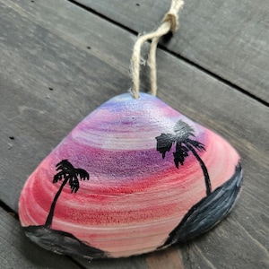May include: Hand-painted seashell ornament featuring a sunset scene with palm trees. The shell is painted with shades of pink, purple, and blue, with black palm tree silhouettes. A natural twine loop is attached for hanging.