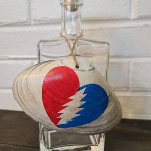 May include: A clear glass bottle with a cork stopper and a white seashell with a red and blue heart painted on it. The heart has a lightning bolt design.