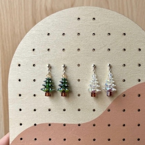 Puede incluir: Dos pares de pendientes colgantes con forma de árbol de Navidad. Un par presenta árboles de cristal facetado verde oscuro con troncos marrones, mientras que el otro par consiste en árboles de cristal facetado transparente e iridiscente.
