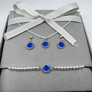 May include: A silver jewellery set comprising a necklace, earrings, and bracelet, each piece set with a bright blue gemstone. The set is displayed in a grey box, tied with a white ribbon, giving a sophisticated, gift-ready look.