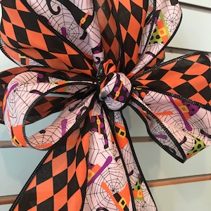 Może przedstawiać: Kokarda halloweenowa z czarno-pomarańczową wstążką w romby i białą wstążką z nadrukiem czarnego kota, kapelusza czarownicy i pajęczyny.