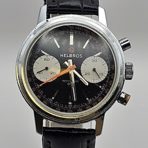 May include: A vintage Helbros Invincible chronograph watch with a black dial, silver case, and black leather strap. The watch face features white and orange accents, with sub-dials at the 6 and 3 o'clock positions.