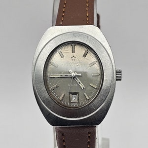 Puede incluir: Un reloj de pulsera plateado con una correa de cuero marrón. La esfera del reloj es plateada con agujas negras y una ventana de fecha que dice "SAHID" y muestra el número 19.