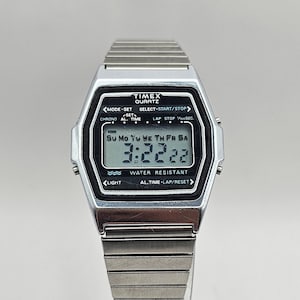 May include: A silver Timex quartz watch with a black digital display. The watch face shows the time, day of the week, and water resistance. The watch has a silver metal band.