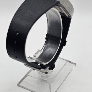 以下が含まれることがあります： 長方形のシルバーケースと黒革バンドのモダンな腕時計。洗練されたデザインを際立たせる透明なアクリルスタンドに展示されています。バンドの幅は約2.5cmです。