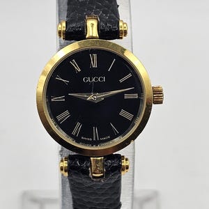 May include: A gold-toned Gucci wristwatch with a black dial and Roman numeral hour markers. The watch features a black leather band and is marked "Swiss Made".